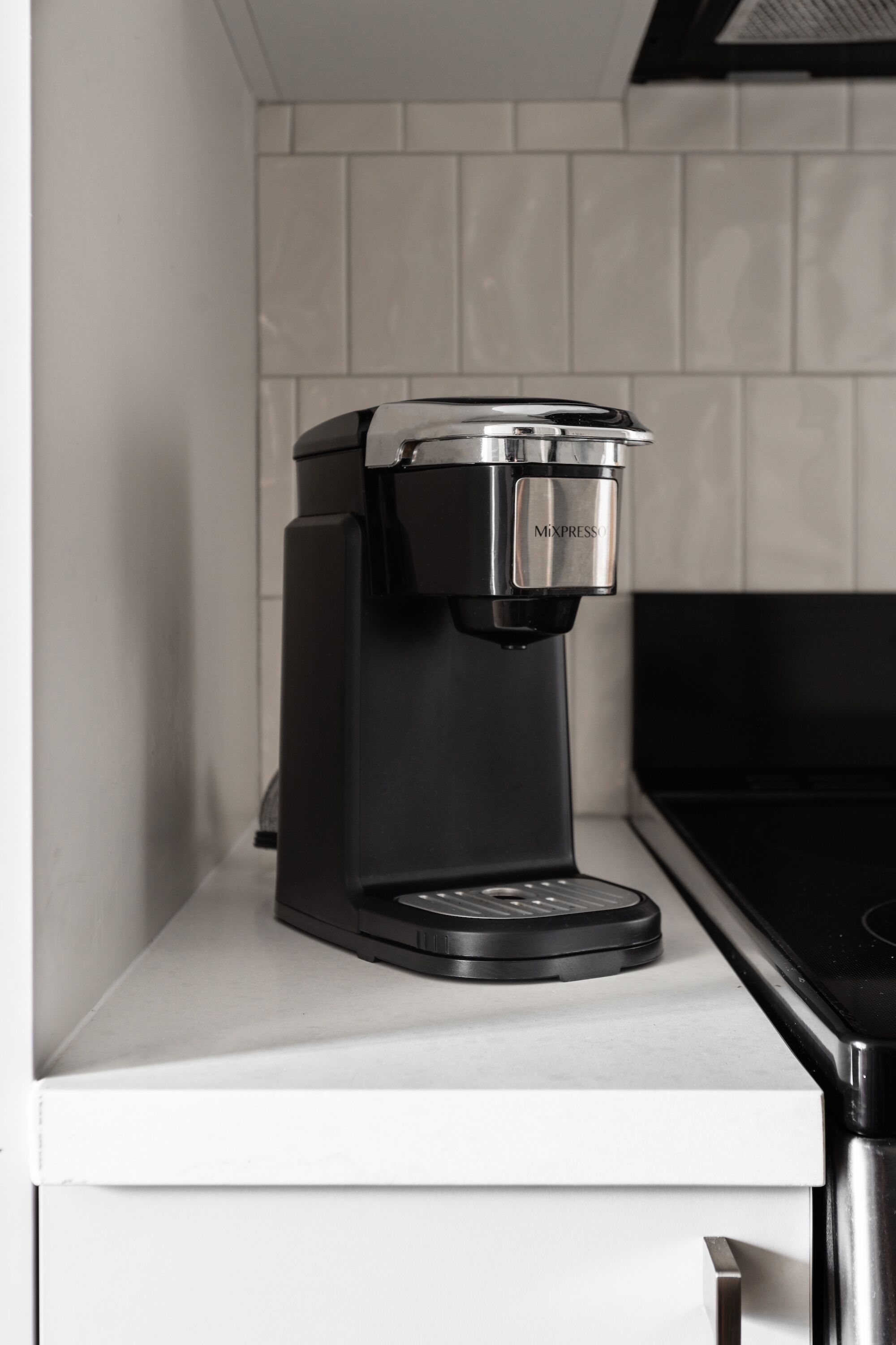 En svart kaffebryggare står på en vit bänkskiva med ett kaklat stänkskydd bakom.