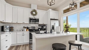A modern kitchen with white cabinetry, a large island with stools, stainless steel appliances, and a view through large windows. - Charming River Townhome A in Gruene (New Braunfels)