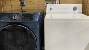 Deux machines à laver, une bleue et une blanche, sont placées côte à côte dans une pièce aux murs en bois.