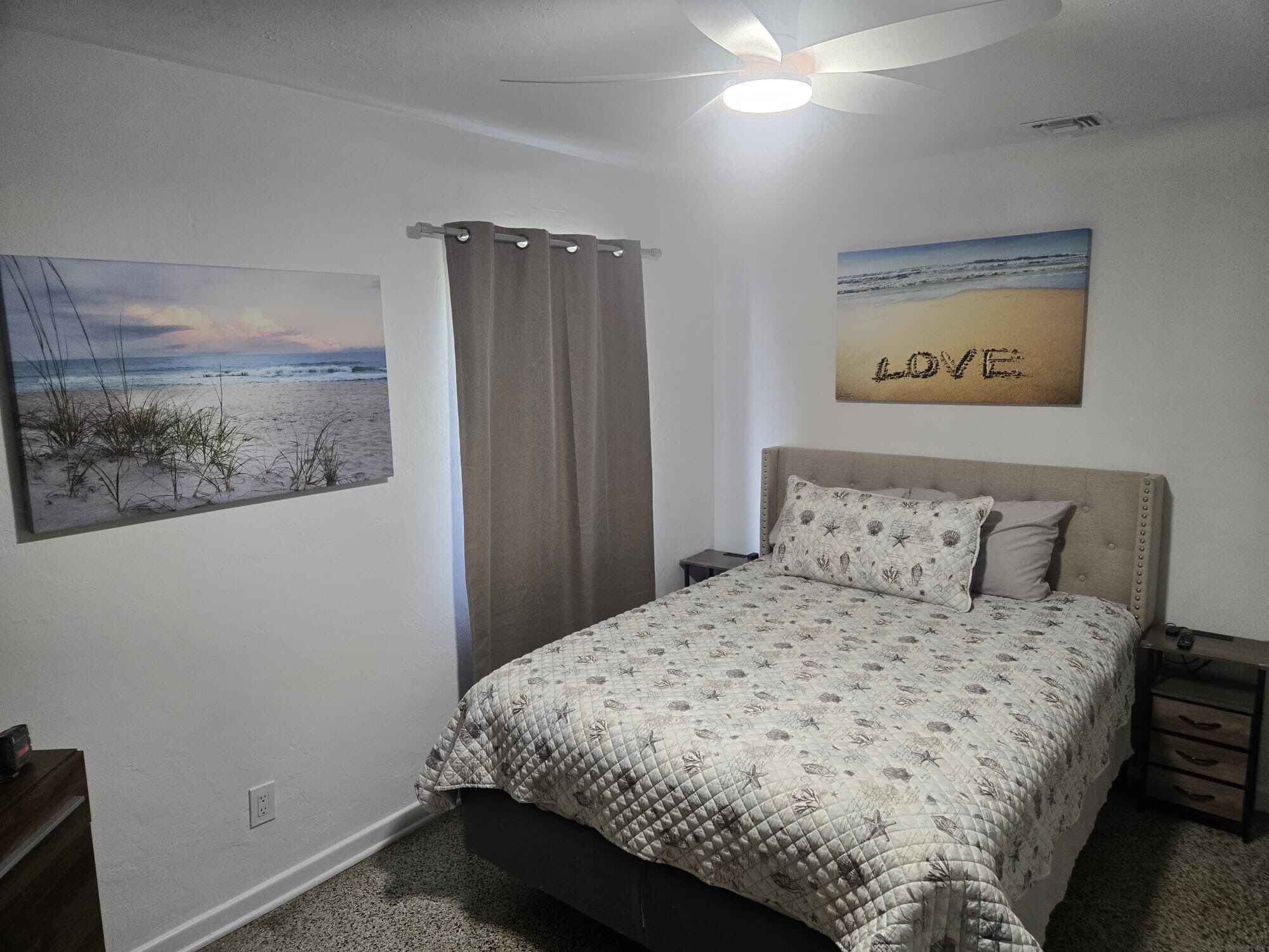 Un dormitorio con cama, ventilador de techo, mesita de noche, escritorio y una pared con una pintura de playa y un letrero que dice 'LOVE'.