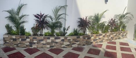 Un patio con suelo de baldosas, una jardinera de piedra con diversas plantas y una pared blanca.
