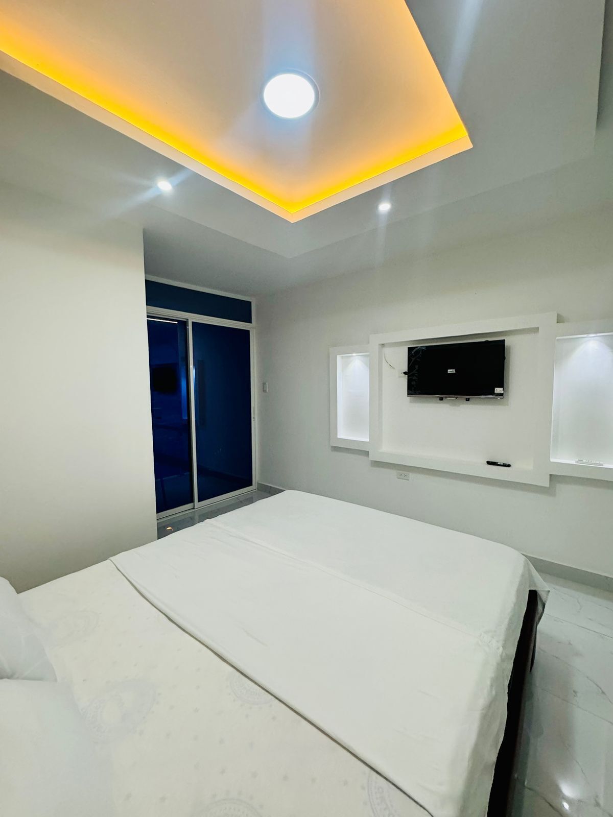 A modern bedroom with a bed, a TV mounted on the wall, and a ceiling light.