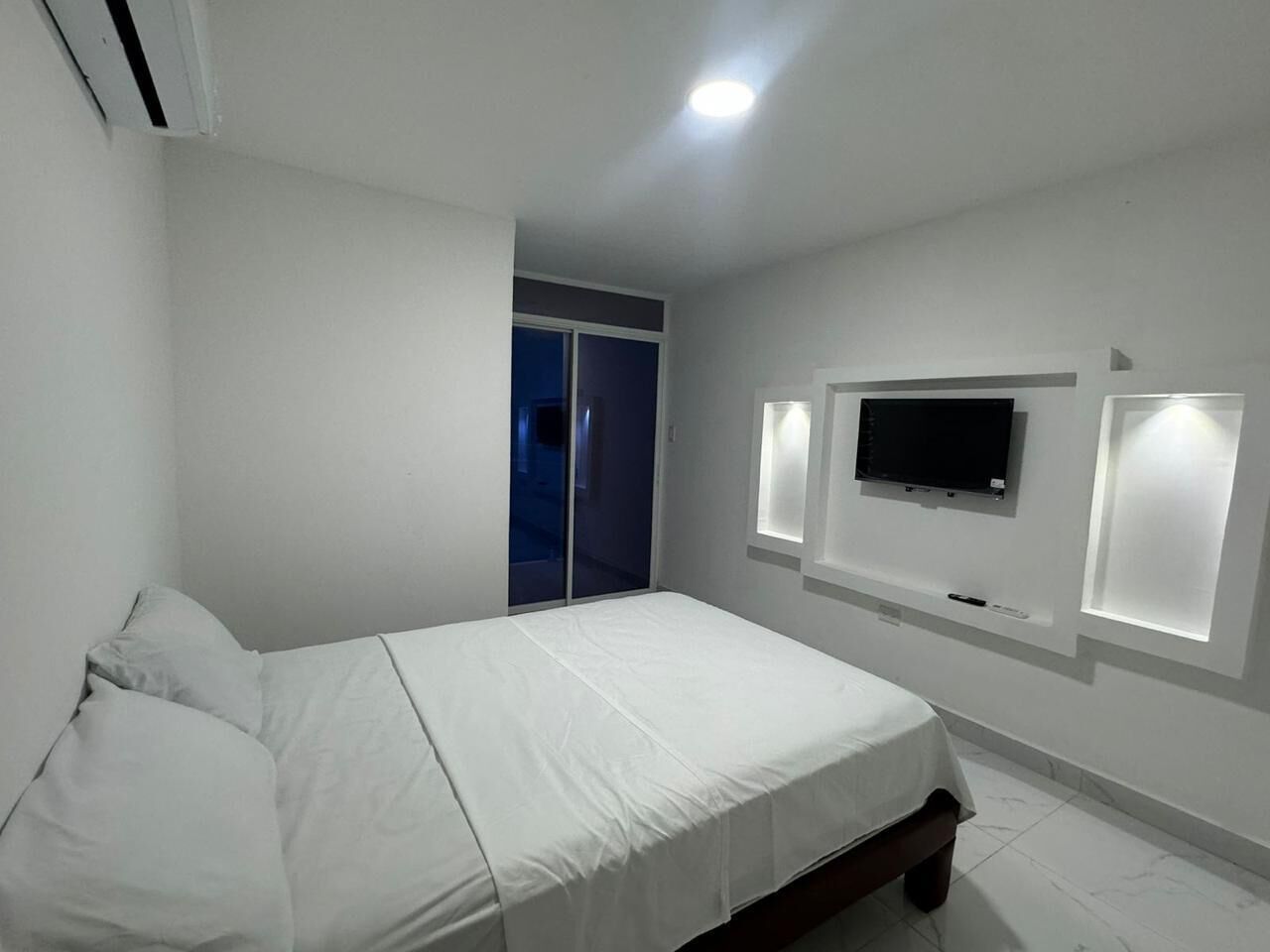 Ein Schlafzimmer mit einem Bett, einem an der Wand montierten Fernseher und einer Schiebetür zu einem anderen Raum.