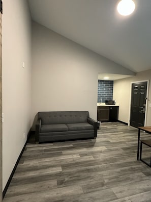 A modern living room with a grey sofa, a wooden floor, and a kitchen area in the background.