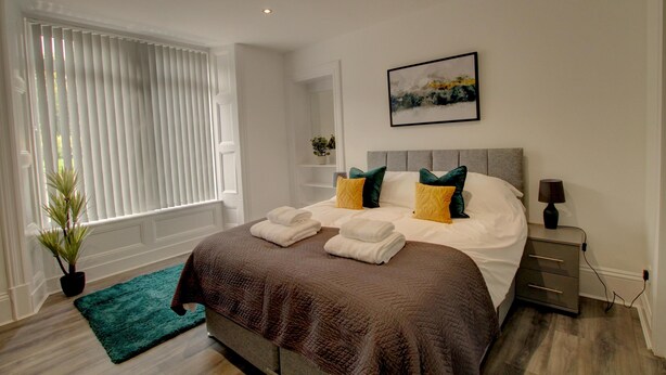 A neatly made bed with pillows, a bedside table with a lamp, and a window with blinds. - Bright & Stylish 1-bed Apt W/free Parking & Wi-fi (Dundee)