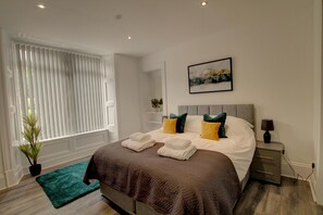 A neatly made bed with pillows, a bedside table with a lamp, and a window with blinds. - Bright & Stylish 1-bed Apt W/free Parking & Wi-fi (Dundee)