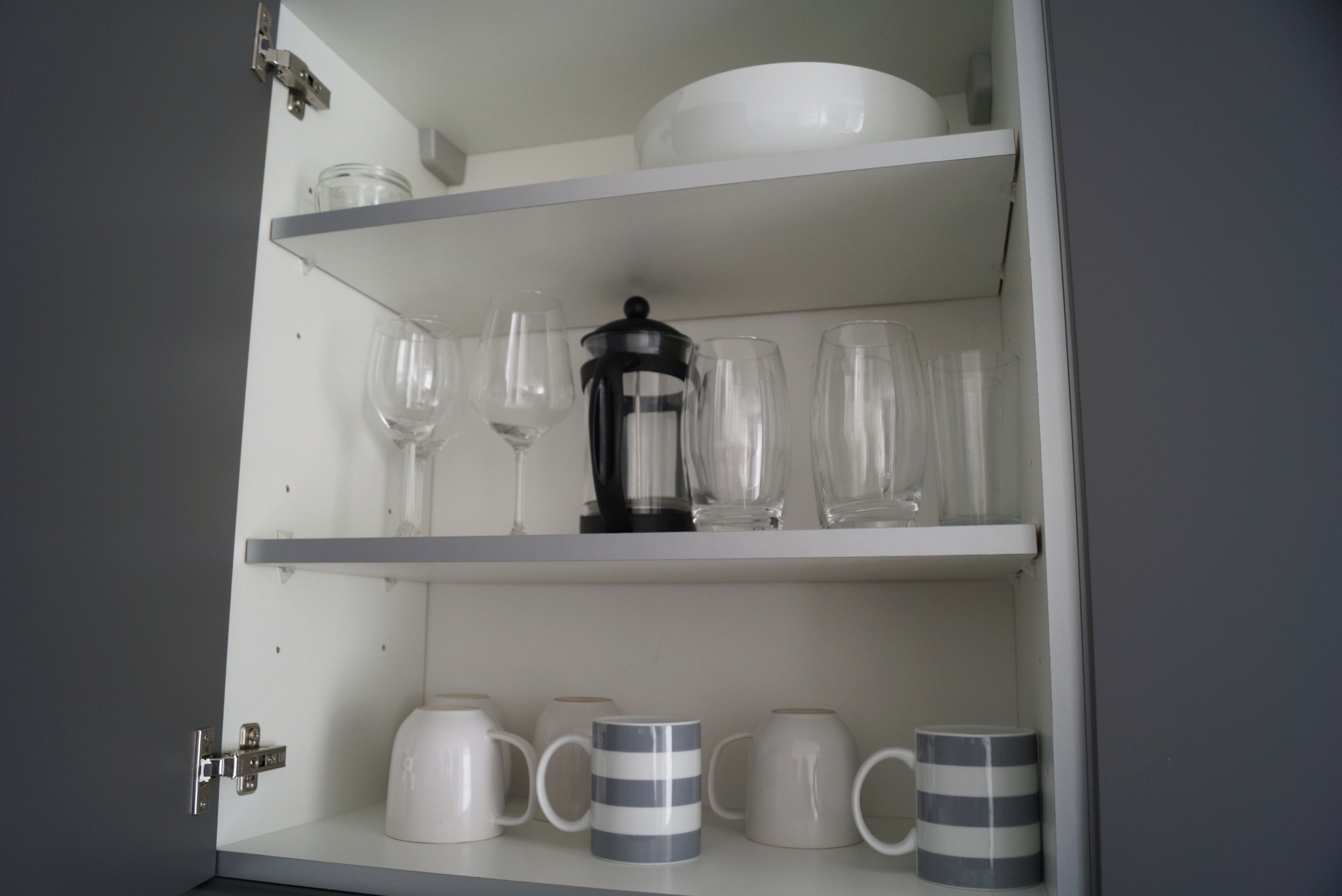Un armario blanco con estantes que contienen cristalería, una cafetera y tazas a rayas.