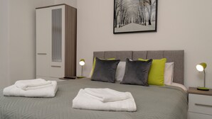 Ein ordentlich bezogenes Bett mit gefalteten weißen Handtüchern, einem grauen Kopfteil und einem gerahmten Bild einer verschneiten Straße an der Wand.