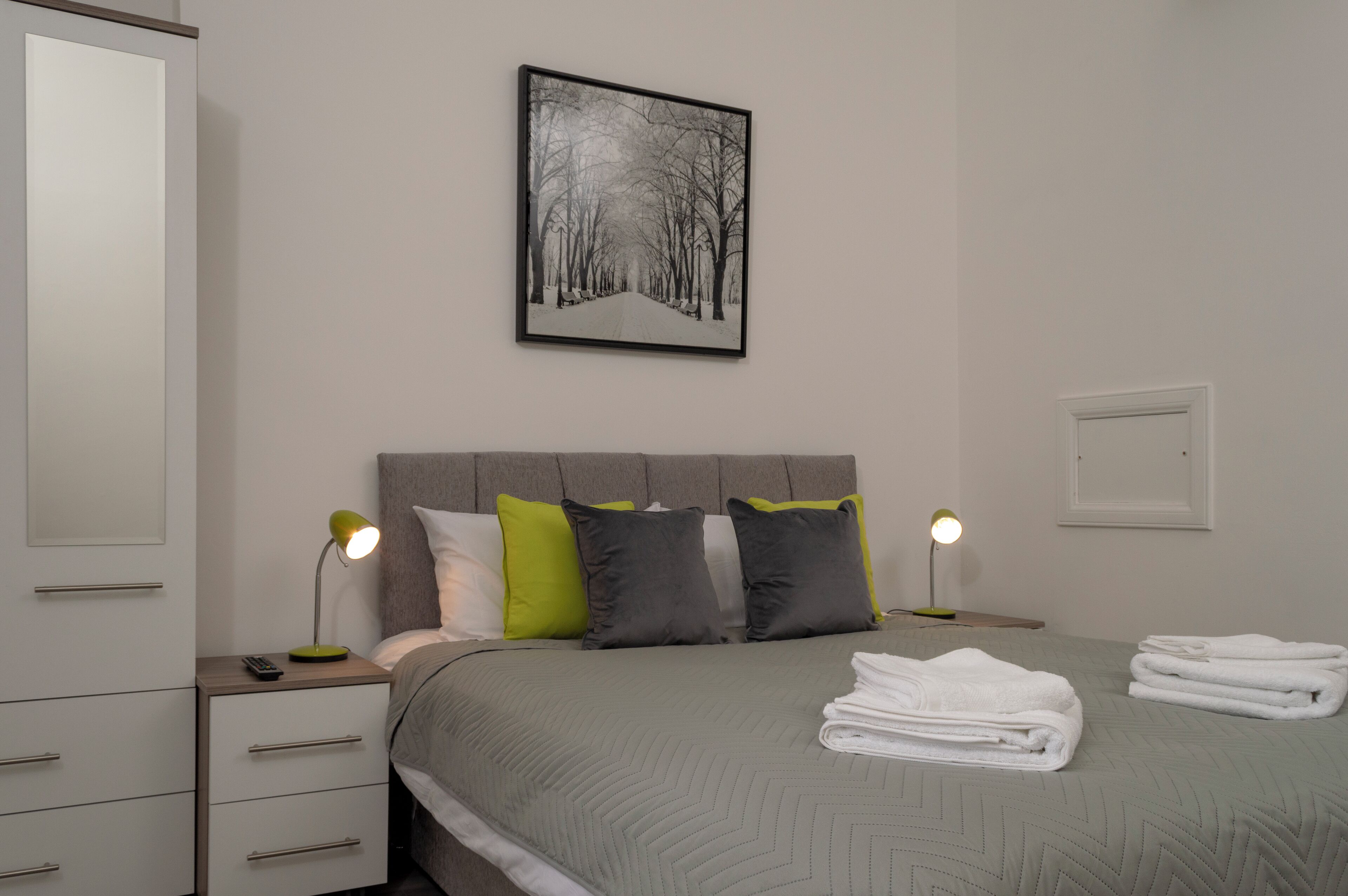 A neatly arranged bedroom with a bed, pillows, bedside lamps, a nightstand, and a framed picture on the wall.