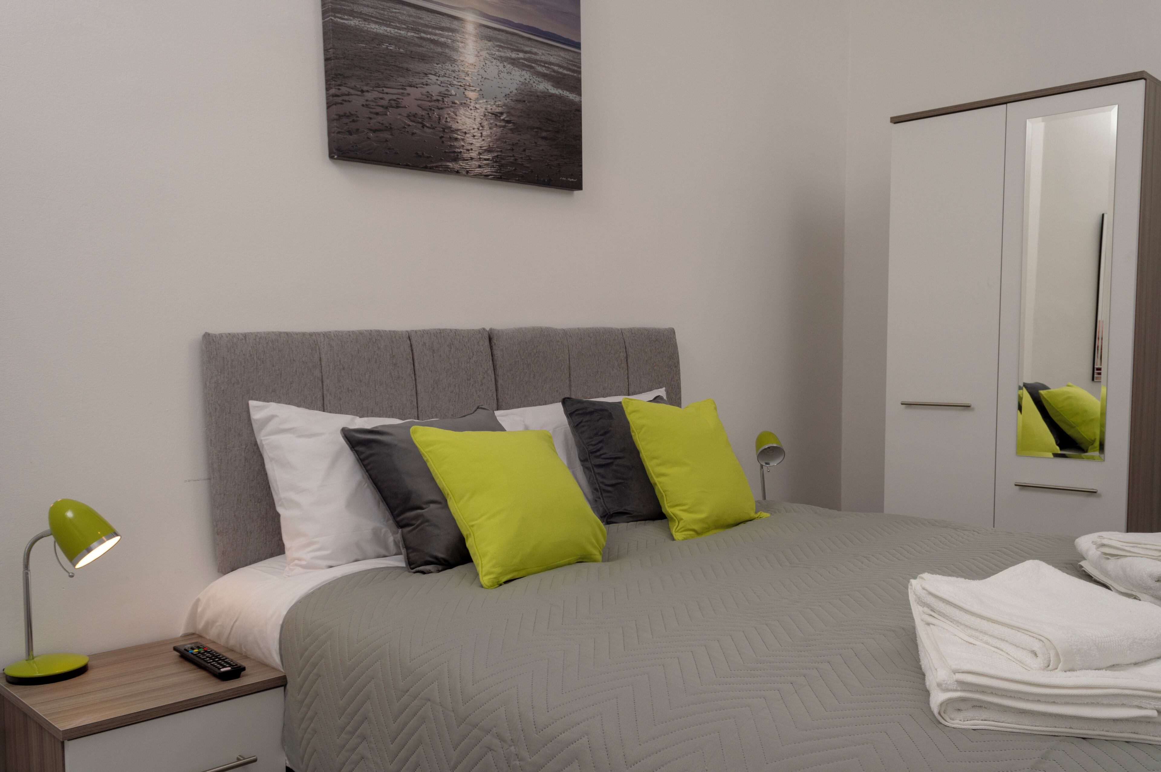 A neatly made bed with a grey headboard, white and green pillows, and a grey quilt. A green lamp is on the nightstand beside the bed. A folded white towel is on the floor next to the bed.