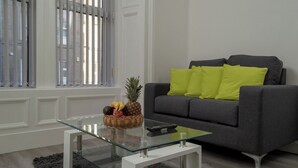 Ein modernes Wohnzimmer mit einem grauen Sofa, einem Glastisch und einem Korbwannen mit Früchten.
