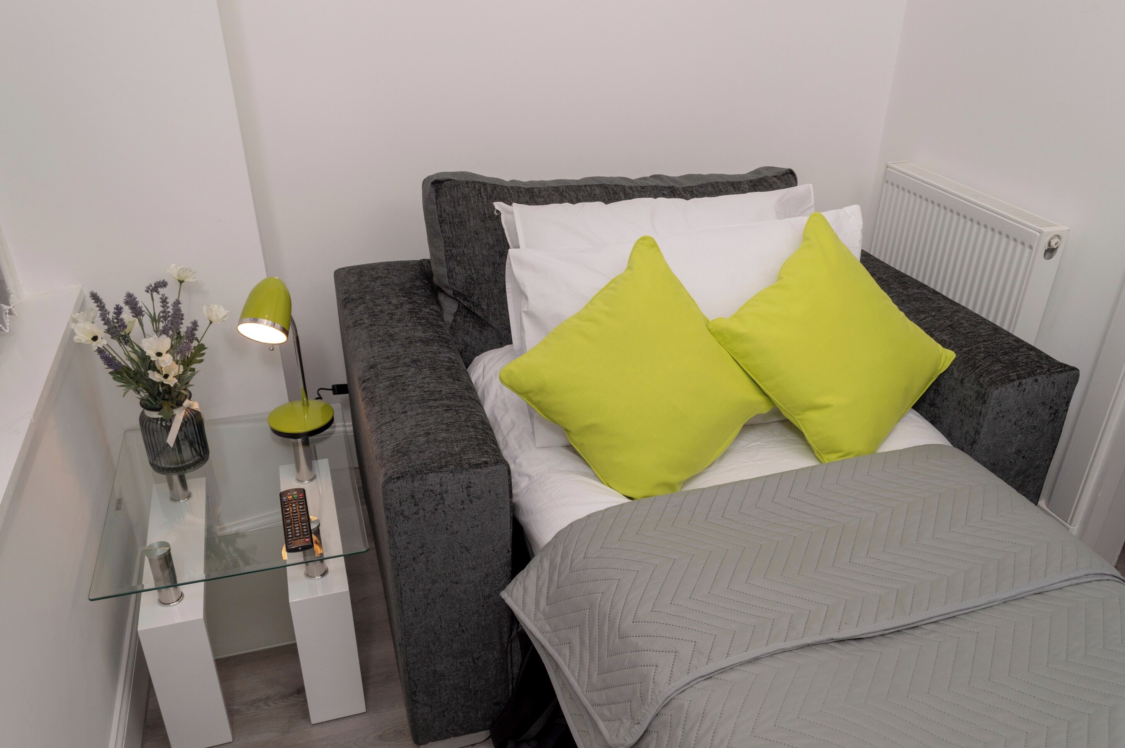 A compact bed with grey bedding and two bright green pillows, a small bedside table with a lamp, and a vase with flowers.