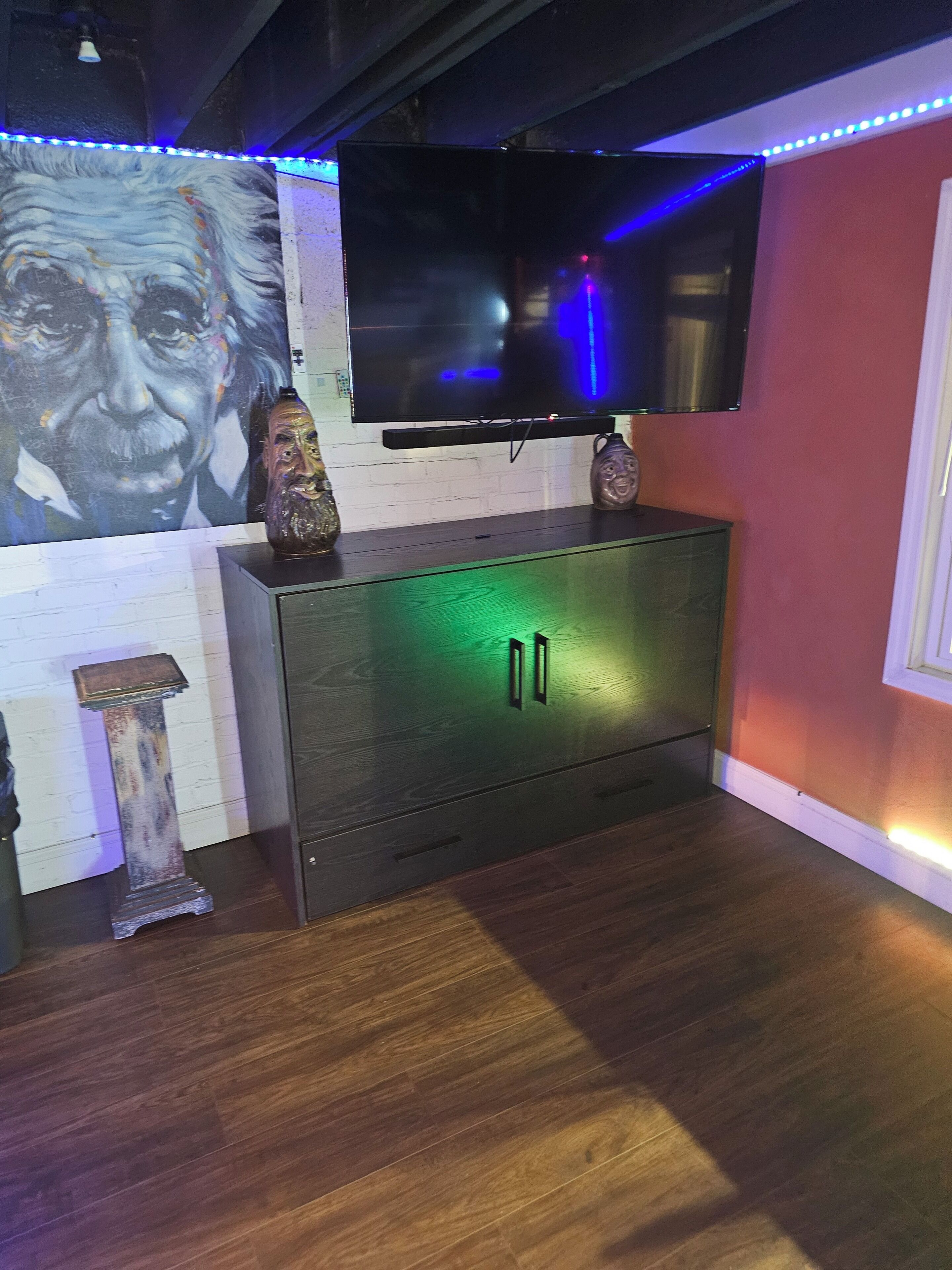 Une pièce dotée d’un téléviseur à écran plat fixé au mur, d’un meuble en bois avec une lumière verte, d’un vase décoratif et d’un grand portrait.