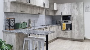 A modern kitchen with grey cabinets, a marble countertop, and a built-in oven. There are two white stools at the island and a potted plant on the left. - Sunset Bliss Paradise in Lilongwe (Lilongwe)