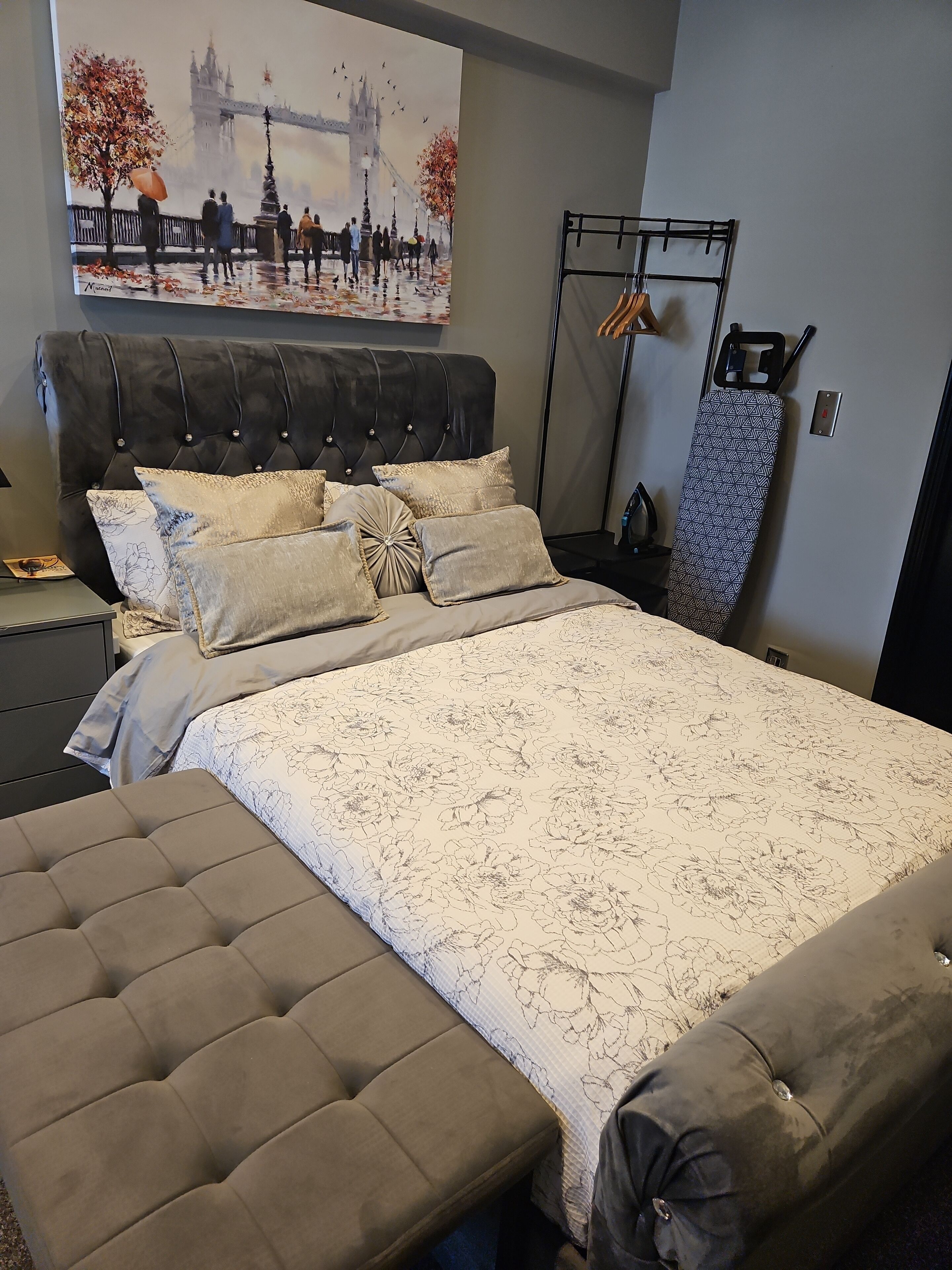 A bedroom with a large bed, a tufted bench, and a painting of a bridge.