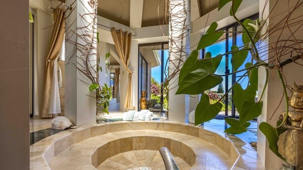 Un área de piscina interior lujosa con un jacuzzi circular, rodeada de suelo de mármol, amplias ventanas y plantas tropicales.