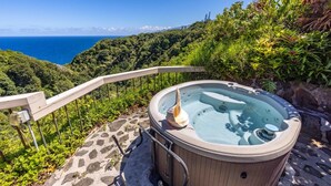 Jacuzzi sobre una terraza de piedra con barandilla, con vista a un paisaje verde y exuberante y al océano.