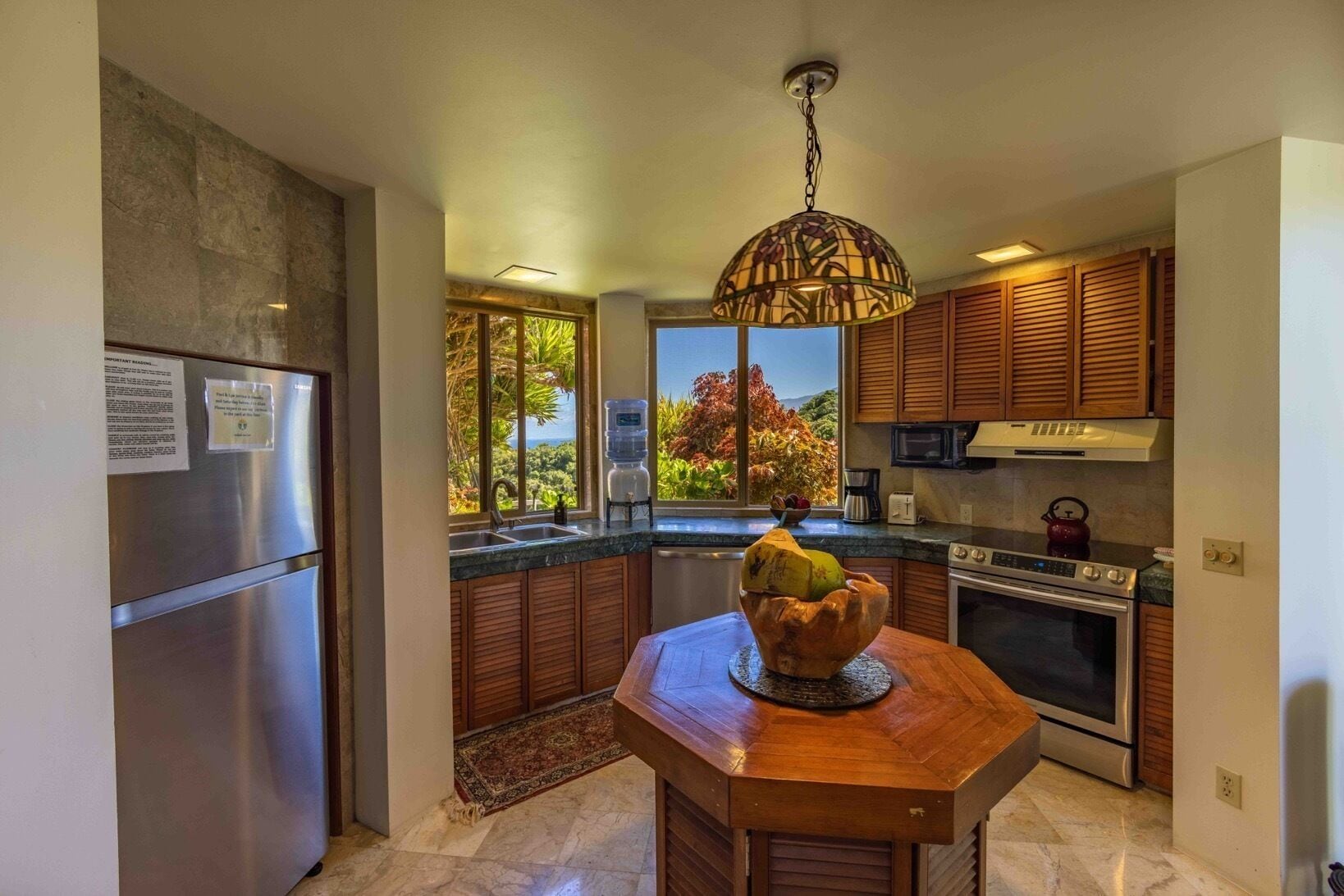 Cocina moderna con una isla central, electrodomésticos de acero inoxidable, armarios de madera y vistas a vegetación exuberante.