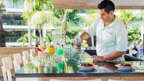 A person preparing drinks at a bar with a tropical view, including palm trees and a building in the background. - Luxury Waterfront 2BR Condo w/ Pool, Gym & Bar, Puerto Vallarta (Puerto Vallarta)