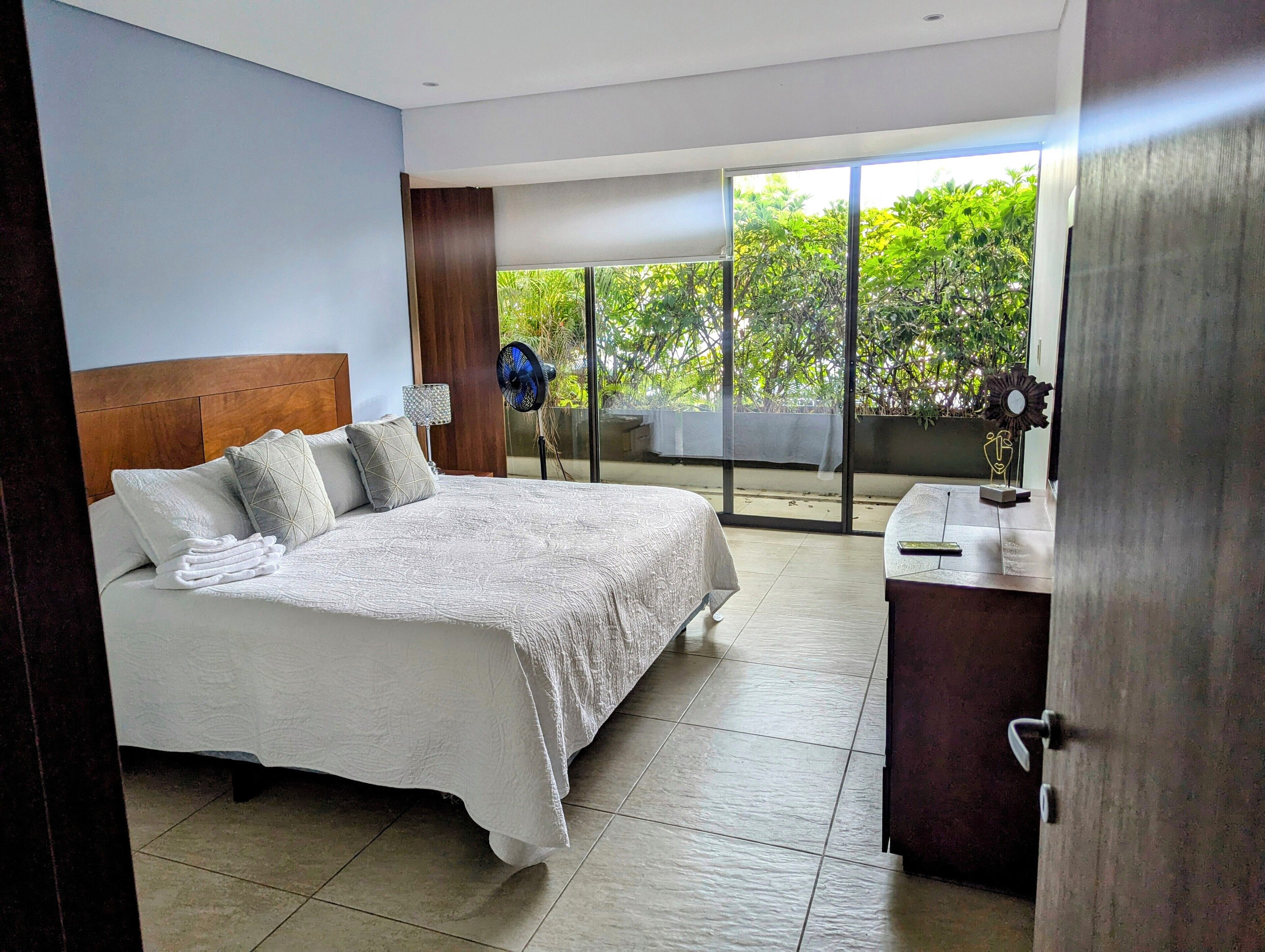 A bedroom with a large bed, a nightstand, a fan, and a view of greenery outside.