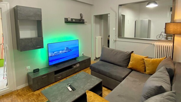 A modern living room with a grey sofa, a flat-screen TV, a coffee table, and a ceiling light fixture. - Sweety flat Marina Zeas Piraeus nearby the sea (Pireas)