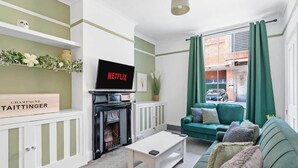 Ein Wohnzimmer mit Kamin, einem grünen Sofa, einem weißen Couchtisch und einem Fernseher, der 'NETFLIX' anzeigt.