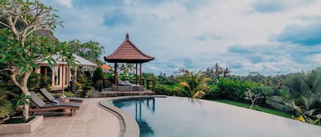 Una piscina infinita tranquila con un pabellón y sillas de descanso, rodeada de exuberante vegetación y un cielo despejado.