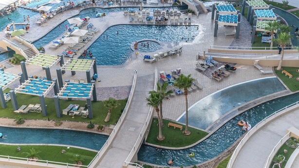 A large outdoor swimming pool area with multiple sections, including a hot tub, surrounded by lounge chairs and umbrellas.