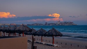 A beach with thatched umbrellas and lounge chairs, a clear view of the sea, and a distant cityscape with buildings and a hill.