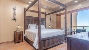 A four-poster bed with a wooden frame, a bedside table with a lamp, and a view of the sea through a balcony.