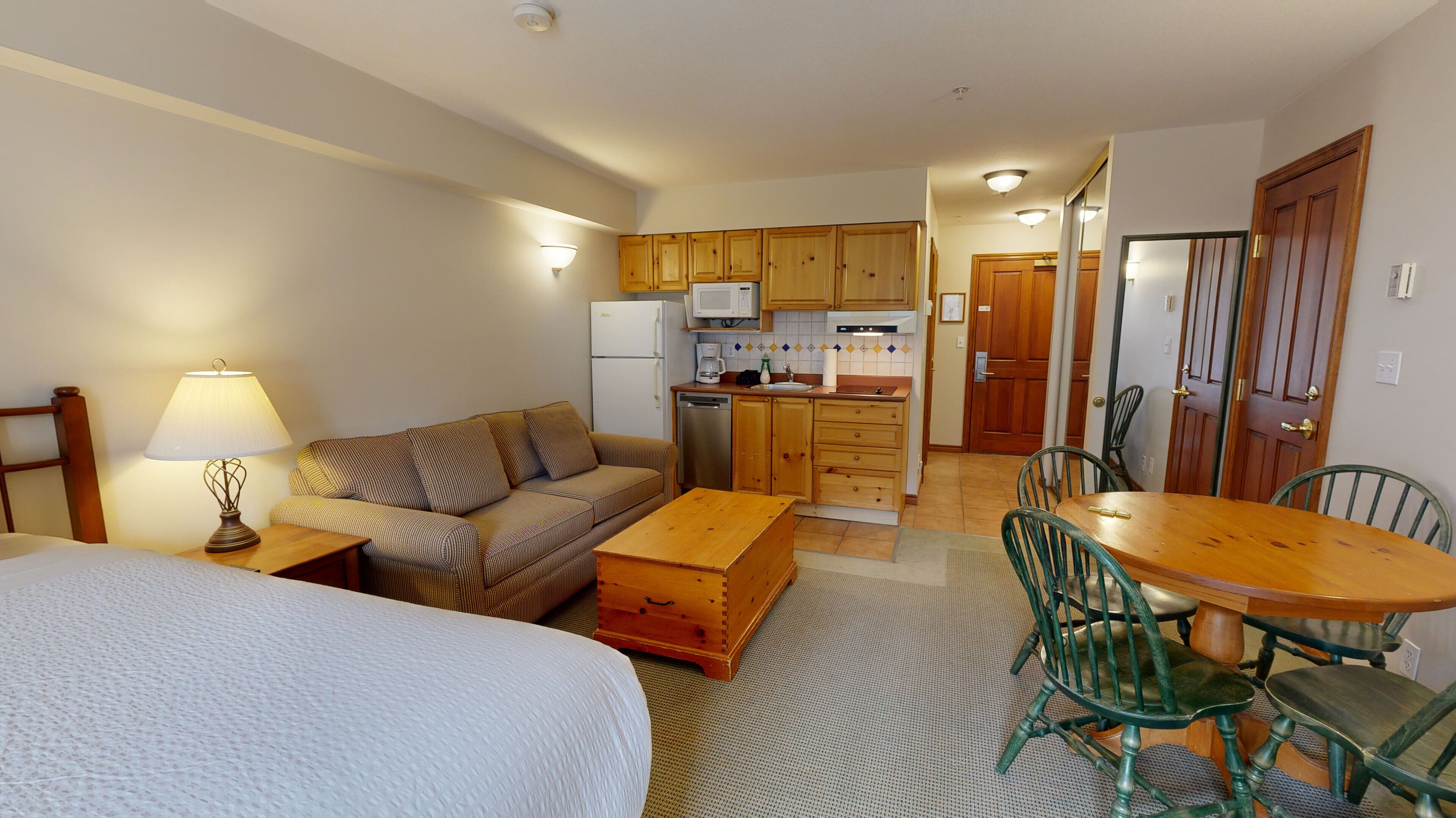 A compact hotel room with a bed, a sofa, a wooden dining table with chairs, a kitchenette, and a small living area.
