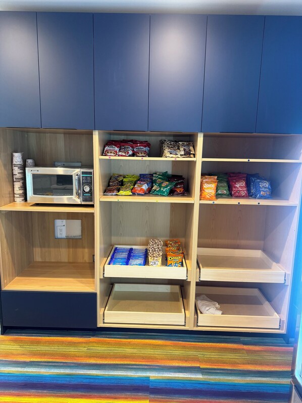 Una kitchenette con un microondas, varios paquetes de comida y un cajón con objetos azules.