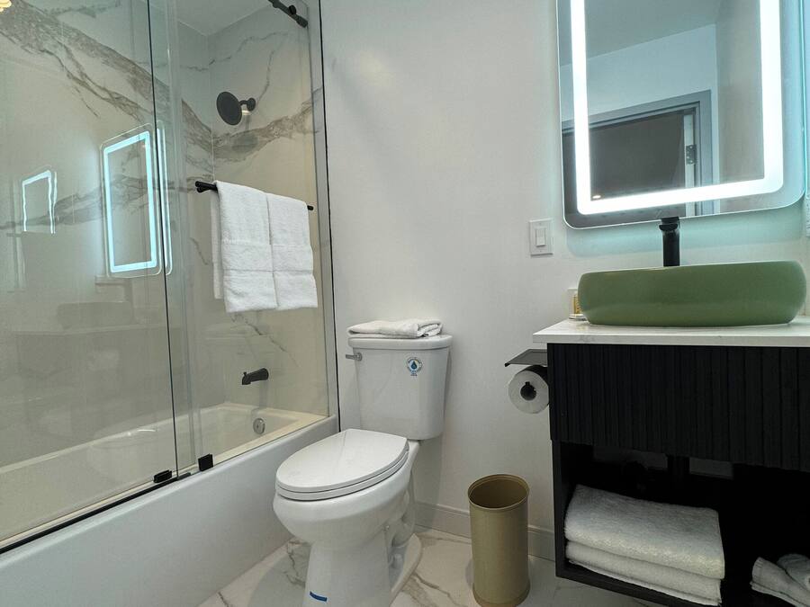 Un baño moderno con ducha envuelta en vidrio, inodoro blanco, tocador con lavamanos verde e iluminación LED en el espejo.
