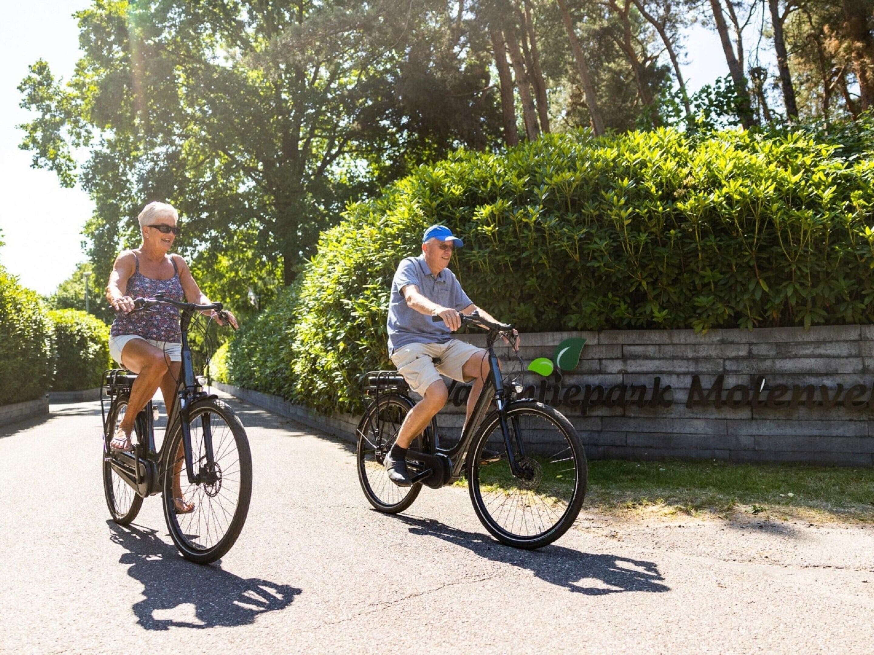 Two people riding bicycles on a paved path with a stone wall and greenery in the background.