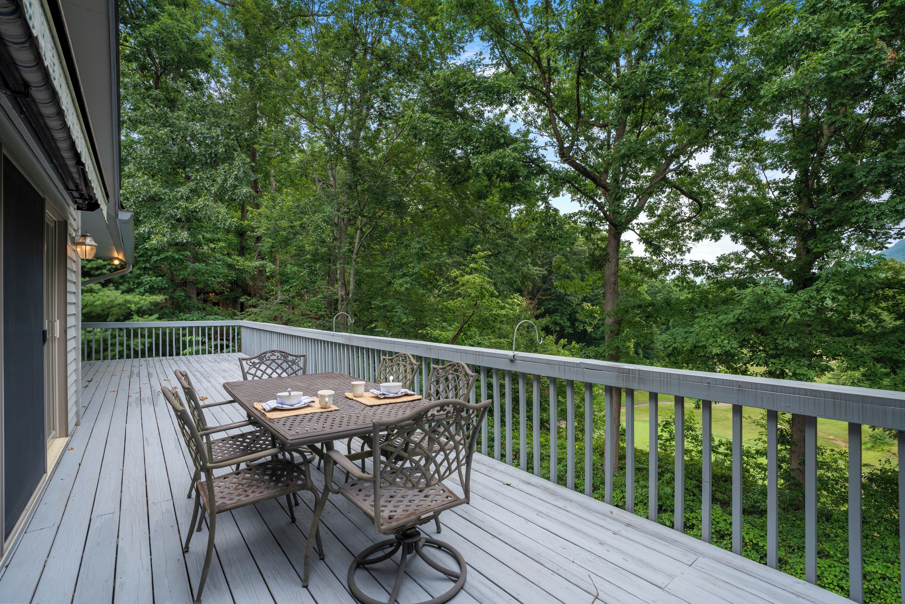Une terrasse en bois avec une table et des chaises, entourée d’arbres et offrant une vue sur un paysage verdoyant.