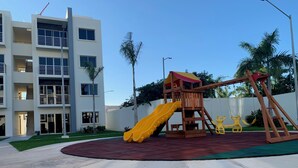 Un terrain de jeux équipé d’un toboggan et de balançoires, entouré d’une pelouse bien entretenue et bordé d’un bâtiment doté de nombreuses fenêtres et de balcons.