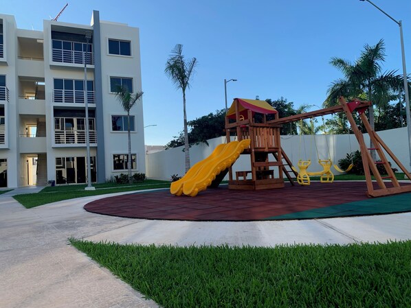 Un terrain de jeux équipé d’un toboggan et de balançoires, entouré d’une pelouse bien entretenue et bordé d’un bâtiment doté de nombreuses fenêtres et de balcons.