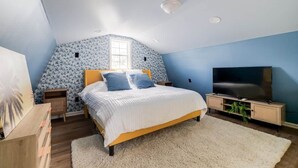 A bedroom with a sloped ceiling, a bed with a white and blue bedspread, a television on a wooden stand, and a window with a view of the outside. - Tiny House 20 Mins from Beach (Brunswick)