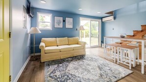 A living room with a blue wall, a beige sofa, a patterned rug, and a wooden table with benches. - Tiny House 20 Mins from Beach (Brunswick)