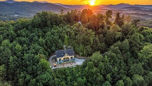 Een lodge omgeven door dicht bos, met uitzicht op een ondergaande zon boven verre bergen.