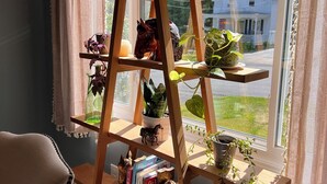 A wooden plant shelf with multiple tiers, holding various potted plants and decorative items, positioned by a window with a view of a house and greenery outside. - The Cardinals Nest, Charming 4-bedroom house, Historic Village of Ballston Spa.  (Ballston Spa)
