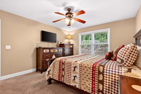 A bedroom with a bed, a ceiling fan, a dresser, a TV, and a window with a view of greenery.