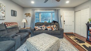 A living room with a grey sofa, a matching ottoman, a ceiling fan, and a large window with curtains. - Beary Cozy | Pets, Work Station & 20 Min. from AVL (Black Mountain)