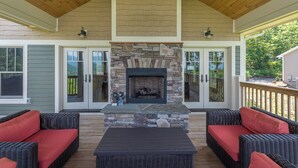 Une terrasse couverte, aménagée avec des meubles en osier, une cheminée en pierre et des portes vitrées donnant sur une terrasse en bois.