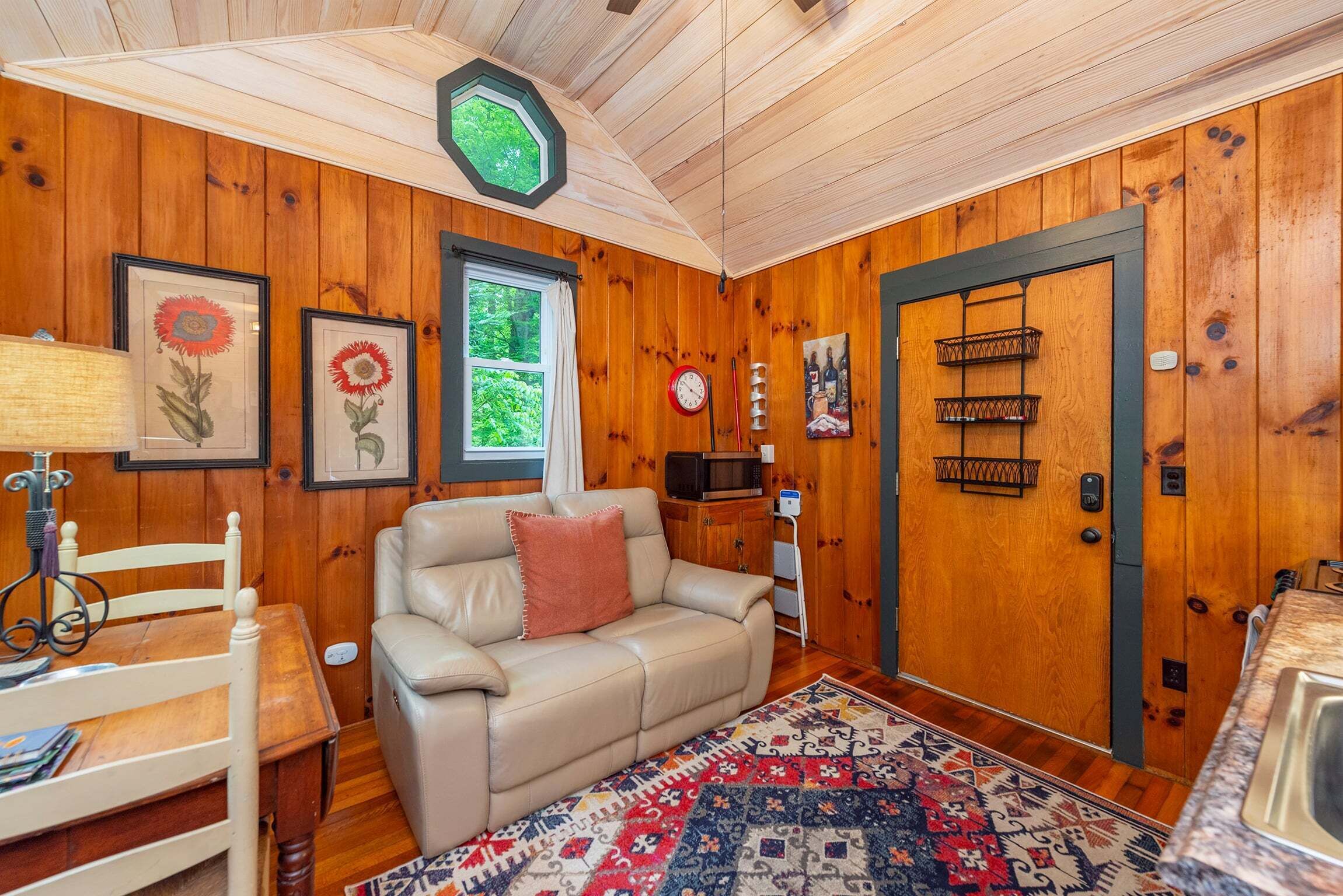 A cozy room with wooden walls, a leather sofa, a patterned rug, a dining table, and a window with curtains.
