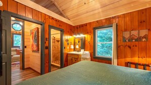 Ein Schlafzimmer mit Holzwandverkleidung, einem Bett, einem Badezimmer mit Waschbecken und Spiegel sowie einem Fenster mit Blick auf Bäume.