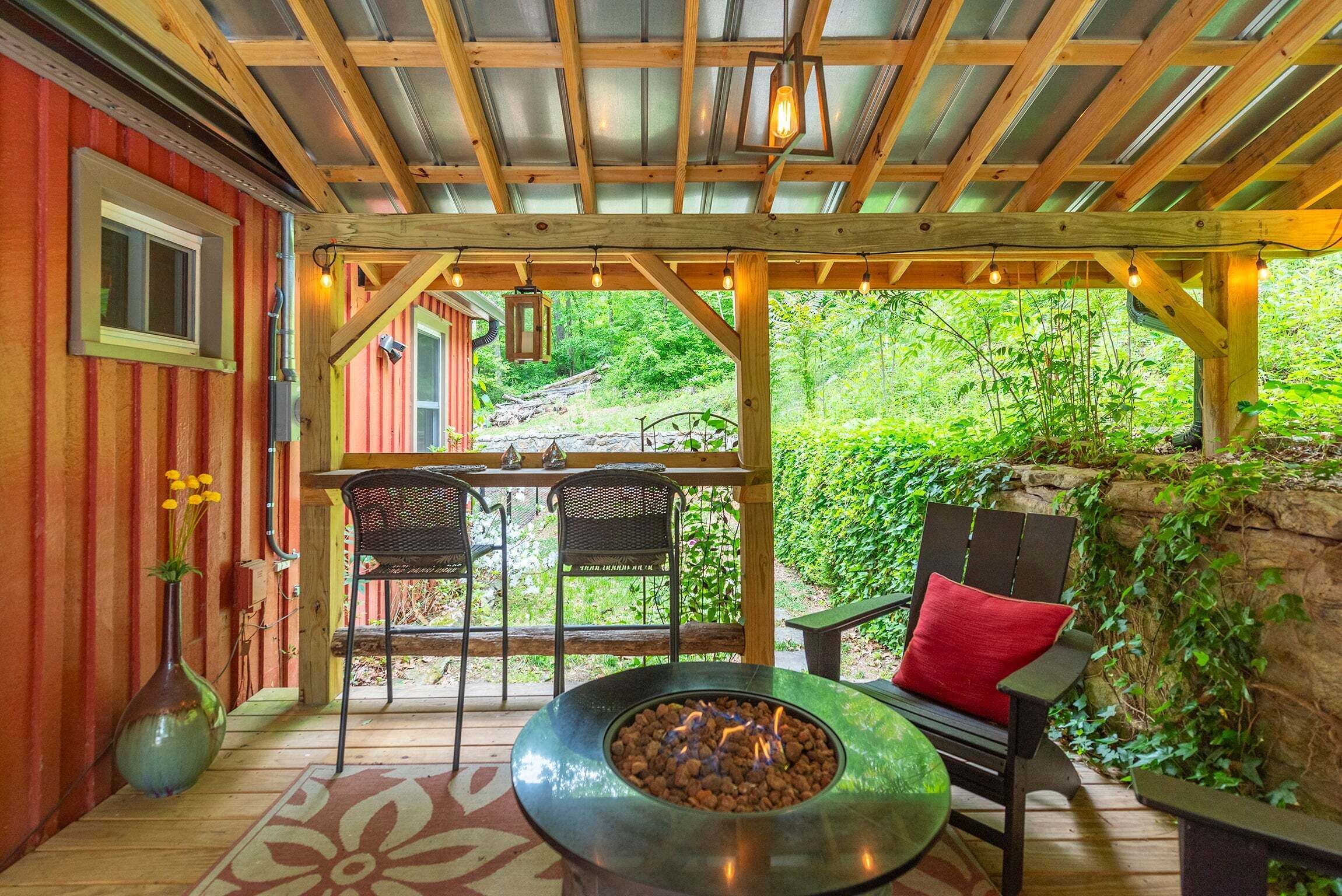 A covered outdoor seating area with a fire pit, wooden tables, chairs, and a view of a garden.