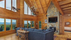 A log cabin interior with a large stone fireplace, a sectional sofa, and a dining area with a wooden table and chairs. - Grandview Lodge | Large 4 Bedroom Mountain Cabin with Hot Tub & Views! (Old Fort)