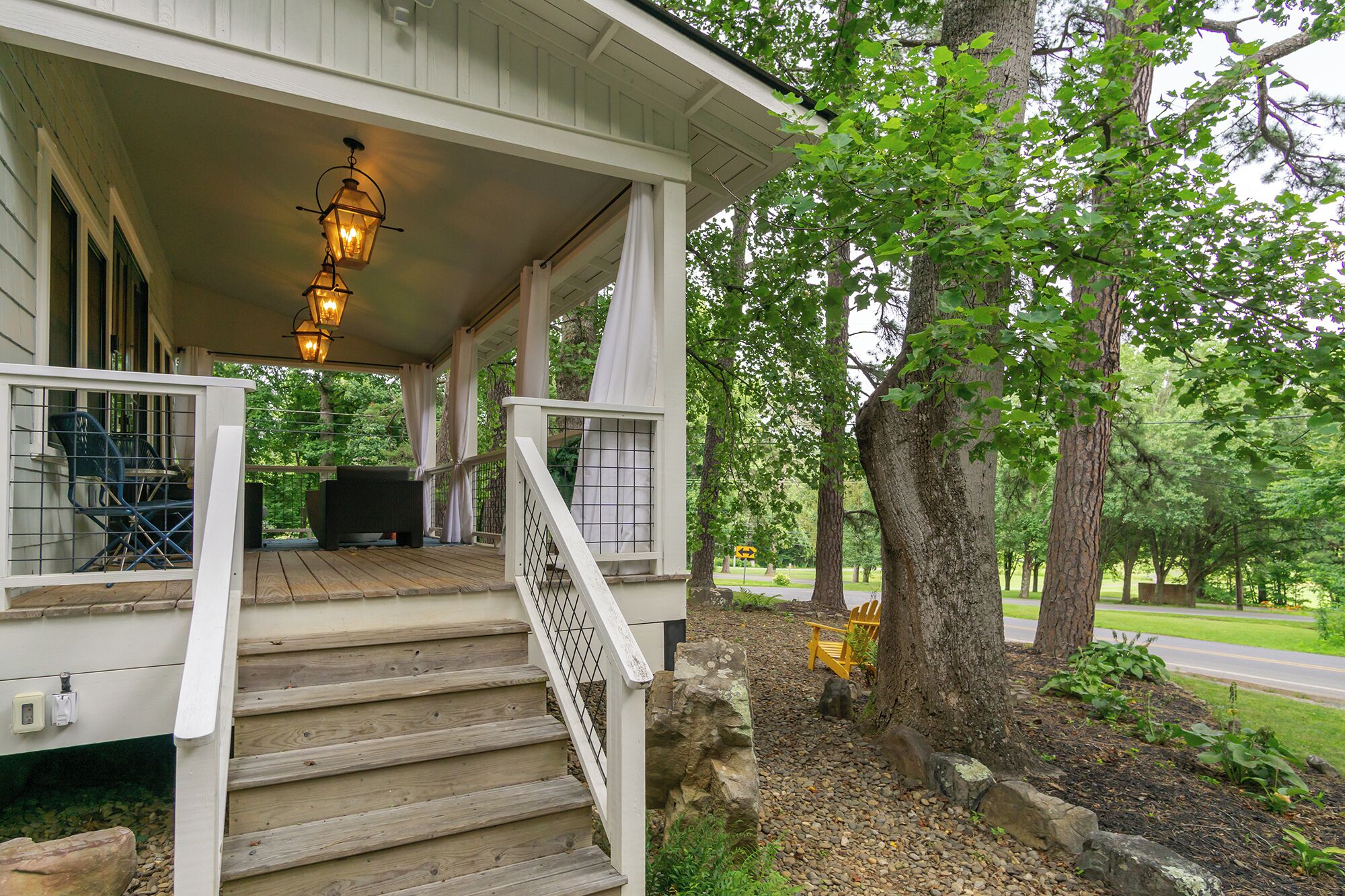 A covered porch with wooden steps leading to a seating area, surrounded by trees and a garden.