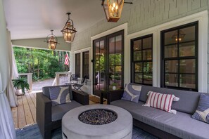 A covered porch with a gray sofa, two chairs, a fire pit, and a view of a forest. - Morning Glory | Luxury Cottage with Covered Porch & Outdoor Fire pit! - 3 Bedroom, 2 Bathroom (Black Mountain)
